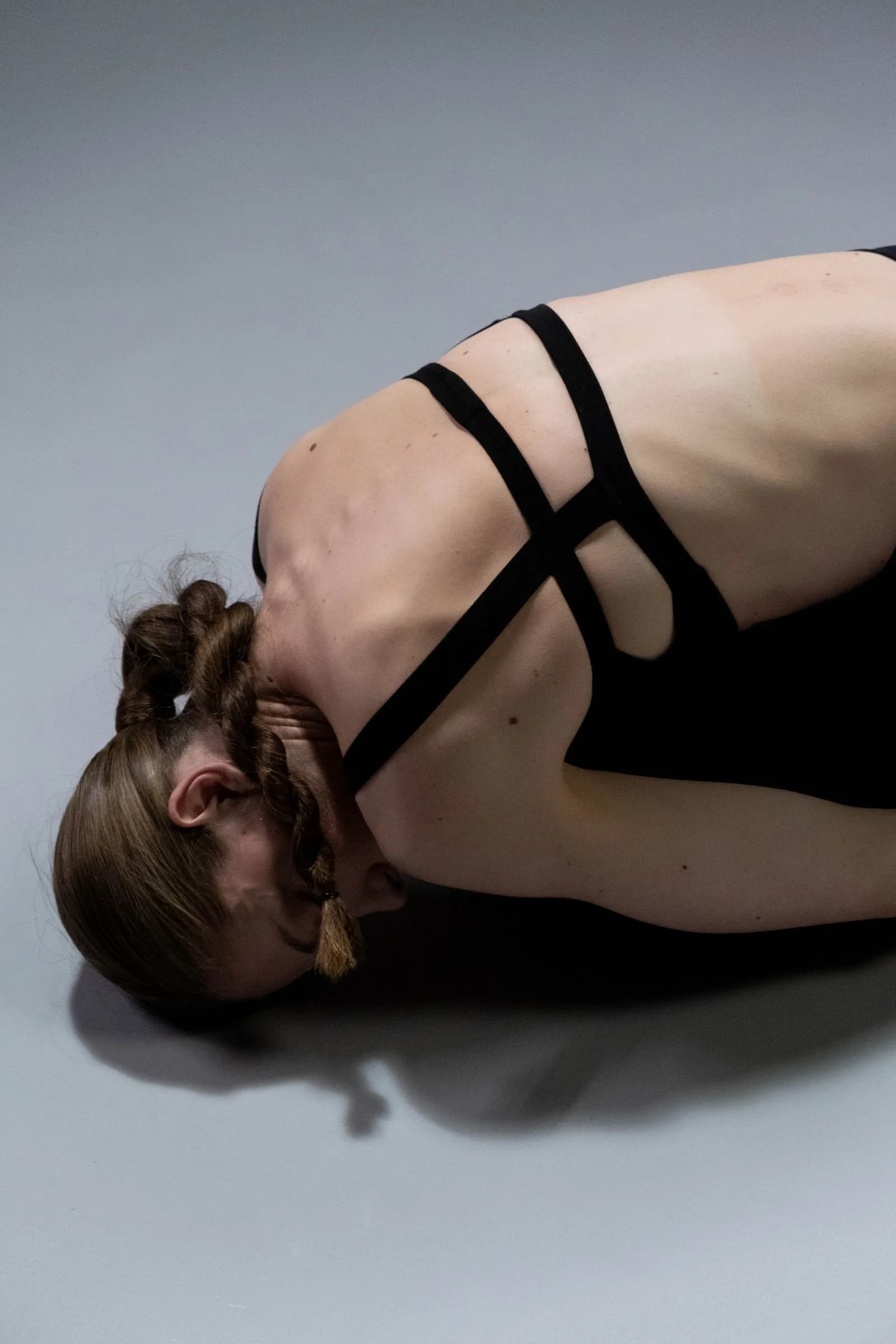Close-up of a woman in a deep forward fold, her head resting between her arms, against a grey studio background.